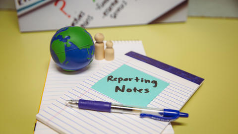 A small globe and set of wooden people rests on top of notepad with a sticky note saying "reporting notes."