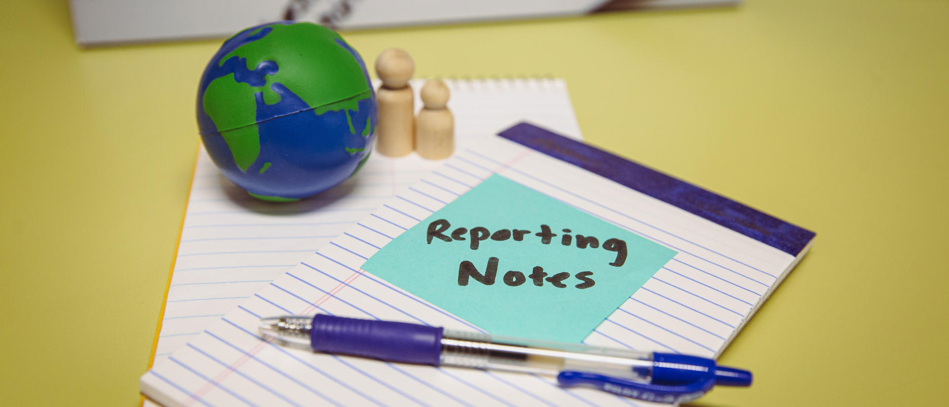 A small globe and set of wooden people rests on top of notepad with a sticky note saying "reporting notes."